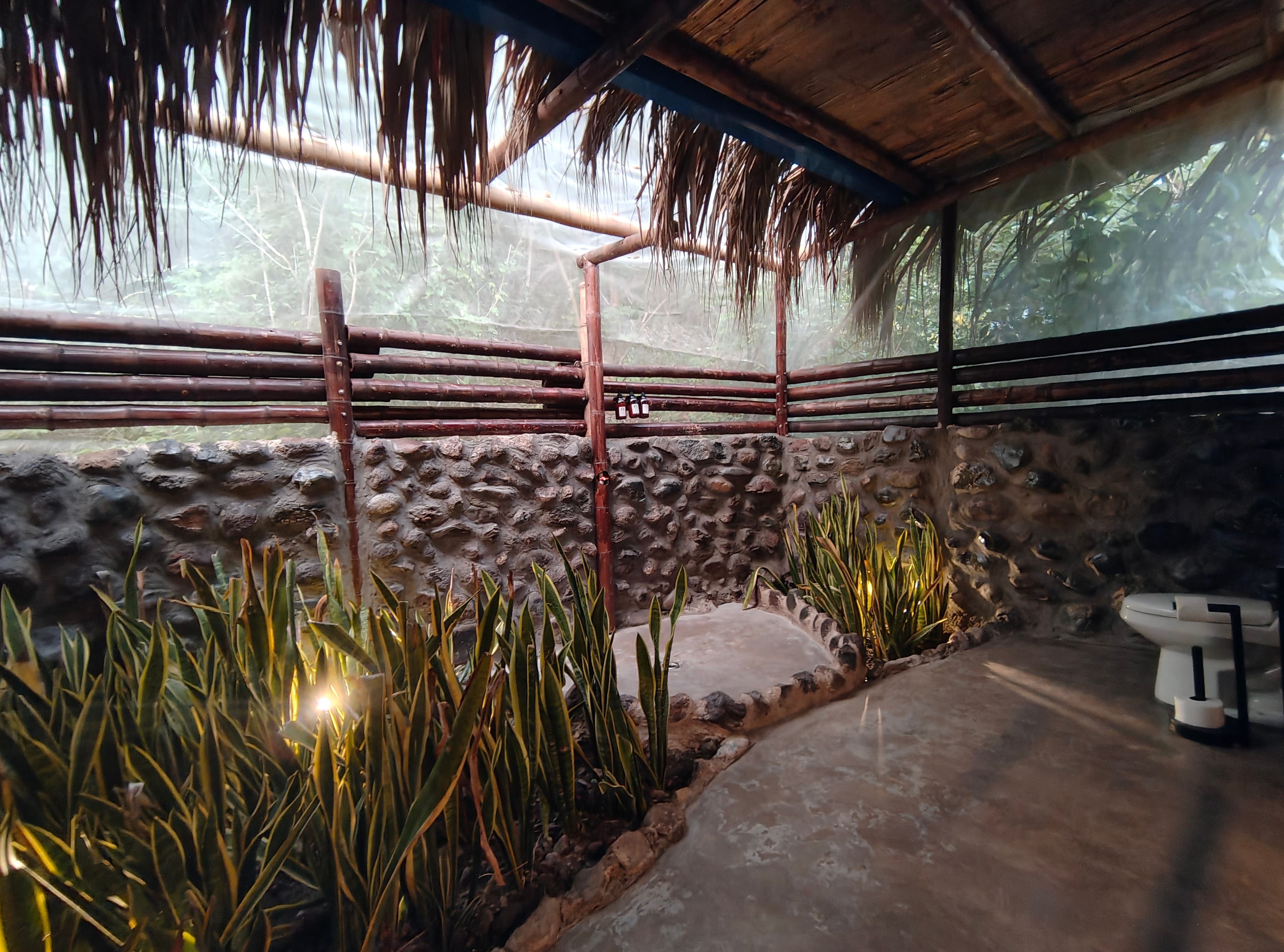 Baño al aire libre con ducha, muro de piedra, plantas tropicales y techo de paja
